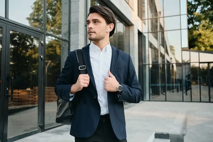 Young handsome bearded brunette man white shirt classic suit with wireless headphones backpack shoulder dreamy looking aside, glass building background
