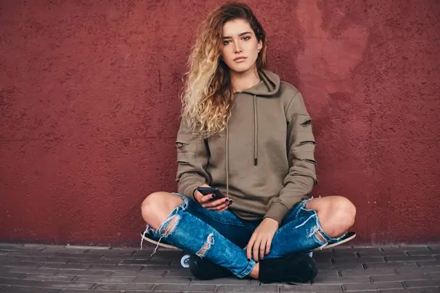 Portrait of young woman in hoodie and ripped jeans, leaning against wall, sitting on skateboard bridge sidewalk