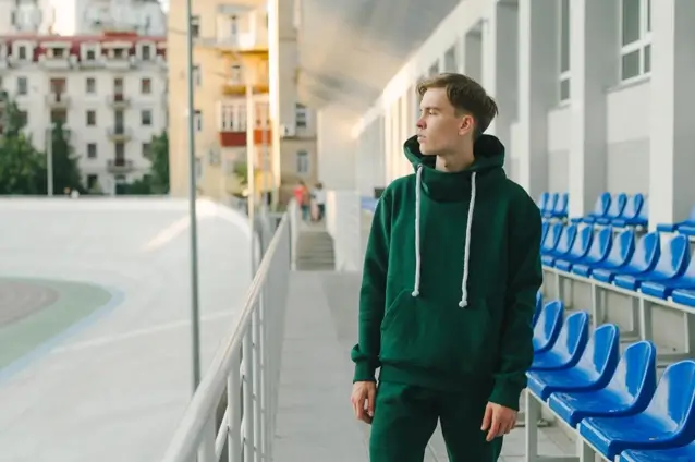 Portrait of a young athletic man in a green hoodie standing near the grandstands at a racetrack looking away with a serious expression