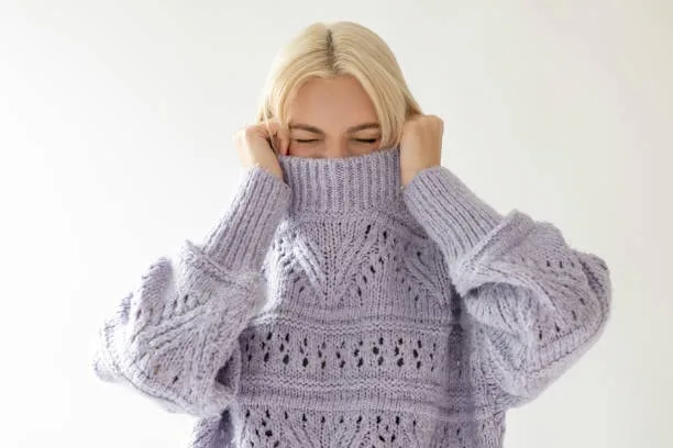 Person wearing a light purple turtleneck sweater with intricate openwork knit patterns, featuring a high folded collar and long sleeves, pulled up to partially cover their face.