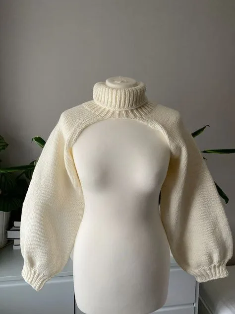 Cream-colored cropped turtleneck sweater with voluminous sleeves displayed on a mannequin in a minimalist setting with a plant in the background.