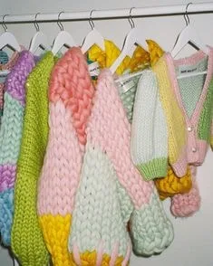 Assortment of colorful chunky knit sweaters on hangers, including pastel color-block designs and a pearl-embellished cardigan, showcasing diverse sweater styles for trendy fashion.