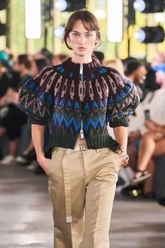 Model on a runway wearing a cropped sweater with a geometric pattern in blue, maroon, and green, featuring a front zipper and balloon sleeves, showcasing bold sweater styles for high fashion.