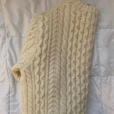Cream cable knit Aran sweater with intricate vertical patterns, laid flat on a quilted surface, showcasing traditional sweater styles for cozy fashion.