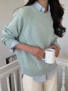 Person in a mint green crew-neck sweater layered over a striped collared shirt, holding a white mug, showcasing types of sweaters for smart-casual layering.