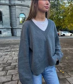 Person wearing a grey V-neck sweater with a deep V-shaped neckline and long sleeves, layered over a white top and paired with light blue jeans, standing outdoors near a building.