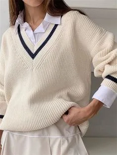 Person wearing a cream V-neck sweater with black trim along the deep V-shaped neckline, layered over a white collared shirt and paired with a white skirt for a preppy style.