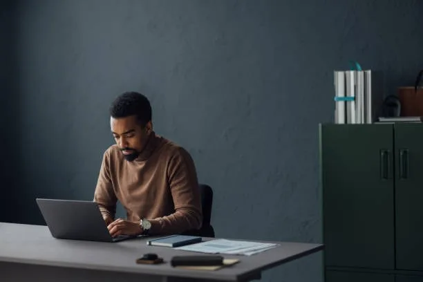 Person wearing a brown turtleneck sweater with a high folded collar, working on a laptop at a desk in a modern office with a green cabinet and documents in the background.
