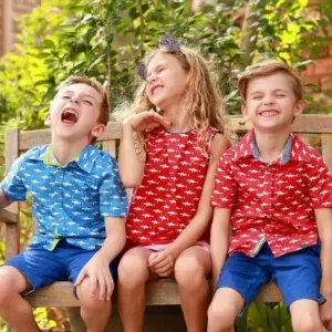 Three children sitting on a bench wearing brightly colored, printed shirts made by a kids wear clothing manufacturer.